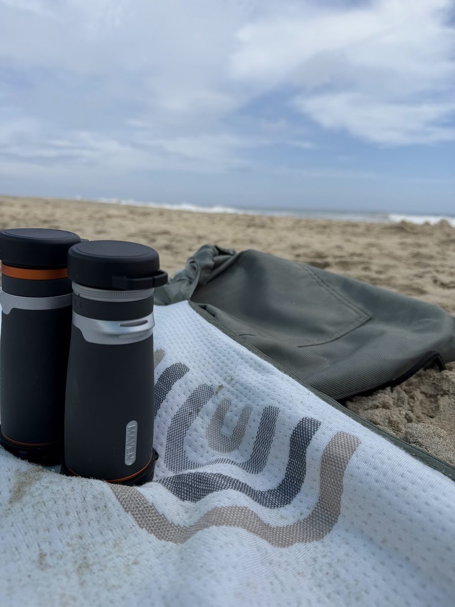 Maven B1.2 10x42 binoculars on a beach towel next to a backpack, with the ocean in the background
